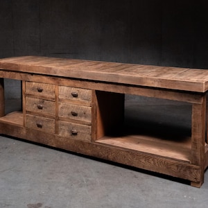 May include: A long, rectangular wooden workbench featuring a flat top surface, a lower shelf, and a central section containing six small drawers arranged in two vertical columns. Each drawer is equipped with a small, dark, circular metal pull.