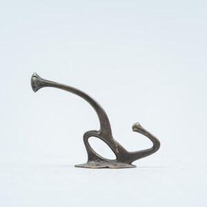 May include: A bronze-toned metal wall hook featuring a dual-prong design with a curved upper arm and a smaller lower hook. The base is flat with two visible screw holes for mounting, and the surface has a weathered, dark metallic finish.