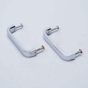Peut inclure: Deux poignées de meuble chromées avec vis. Les poignées sont rectangulaires avec des coins arrondis et une surface brillante. Les vis sont argentées et montrent des signes d'âge. Idéal pour la restauration.