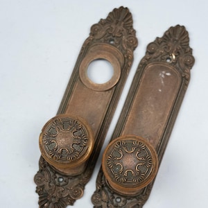 May include: Two antique bronze door handle sets. Each set includes a rectangular backplate with ornate detailing and a round knob with a floral design. The sets are in a vintage style.