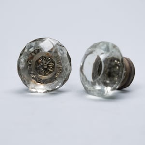 Peut inclure: Deux boutons de porte en verre clair et facetté avec un motif floral sur la base métallique. Les boutons ont une esthétique vintage et sont posés sur un fond blanc. Les boutons sont probablement pour une armoire ou un tiroir.