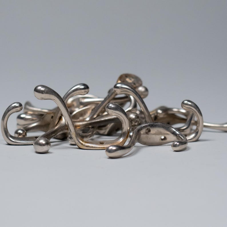 Op de afbeelding: Een verzameling zilverkleurige metalen kapstokhaken. De haken hebben een gebogen ontwerp met een afgeronde bal aan het uiteinde. De haken liggen verspreid op een lichtgrijze ondergrond.