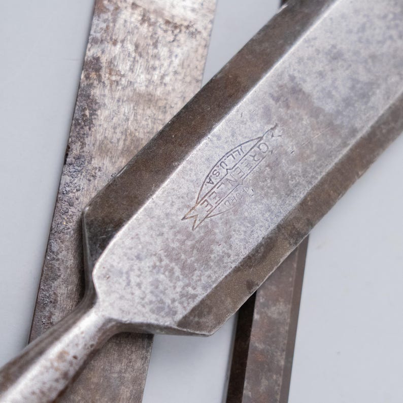 May include: Close-up of vintage metal chisels. The primary chisel features a beveled edge and the text "GREENLEE ILL. USA" stamped on its surface. The tools show signs of wear, with a weathered, metallic appearance, suggesting their use over time.