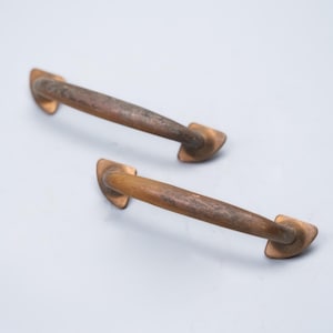 May include: Two antique bronze cabinet pulls. Each pull has a curved handle with a rectangular base on each end. The bronze has a weathered appearance with variations in color, suggesting age and use. The pulls are designed for easy gripping.