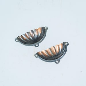 Peut inclure: Deux poignées de tiroir semi-circulaires présentant un motif en forme de coquille festonnée. Chaque poignée possède une finition métallique sombre et patinée avec des reflets cuivrés sur les crêtes et deux trous de fixation.