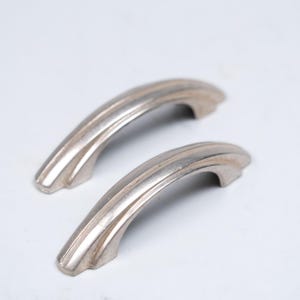 May include: A pair of silver-coloured cabinet handles with a curved, Art Deco design. Each handle has three parallel grooves. The handles are made of metal and are set against a white background. The handles are suitable for furniture.