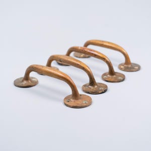 May include: Four antique bronze door handles. Each handle has a curved, arched design with a circular base. The handles show signs of age and wear, with a warm, aged patina. The handles are made of bronze.
