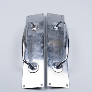 Op de afbeelding: Twee vintage zilveren deurklinken. Elke klink heeft een rechthoekige achterplaat en een gebogen, buisvormige handgreep. Het metaal heeft een gepolijst, reflecterend oppervlak met tekenen van slijtage. De klinken staan tegen een witte achtergrond.