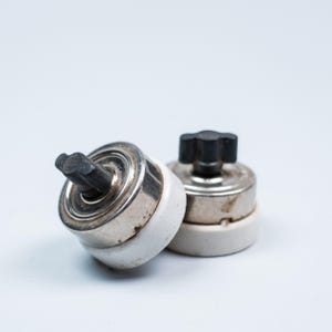 May include: Two circular electrical switches featuring a white ceramic base, a metallic silver-toned middle section, and a black four-pointed knob on top. The switches are positioned close together, showing their cylindrical shape and components.