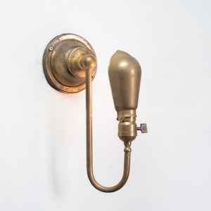 May include: Antique brass wall sconce with a bulb-shaped light fixture. The sconce features a circular backplate, a curved arm, and a bulb holder with a small switch. The fixture is mounted on a white wall.