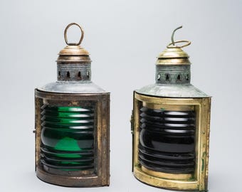 May include: Two antique nautical lanterns. One has a green lens, the other a black lens. Both have a metal frame with a brass top and a handle. The lanterns are designed to be mounted on a ship.