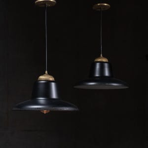 May include: Two pendant lights featuring wide, matte black metal shades with a flared rim. Each fixture has a ribbed, metallic gold-toned cap at the top, connected to a thin black cord and a circular gold-toned ceiling mount against a dark background.