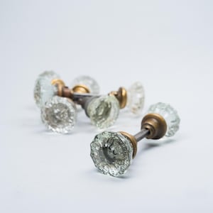 May include: A collection of vintage glass doorknobs with brass hardware. The clear, faceted knobs have a classic, antique appearance. The knobs are arranged on a white surface, showcasing their intricate design and reflective qualities.