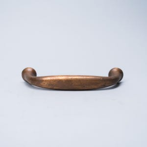 May include: A bronze-colored cabinet pull handle with a curved design. The handle has a smooth, rounded grip and is slightly curved. The ends of the handle are spherical. The finish has a slightly aged appearance.
