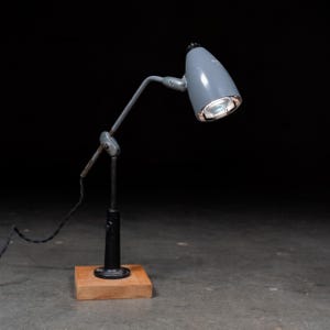 以下が含まれることがあります： グレーの金属製円錐形シェードと調節可能なアームを備えたデスクランプ。ランプは正方形の木製ベースに取り付けられた黒い垂直ポールの上に設置されています。ベースから黒い電源コードが伸びており、ランプヘッドは下向きに角度が付けられています。