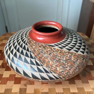 May include: A low-profile ceramic vase with a wide, rounded body and a smooth, solid red rim. The exterior features a contrasting design with one section displaying a dense, speckled brown and tan texture and the other showcasing a geometric pattern of black triangles filled with fine white cross-hatching lines.