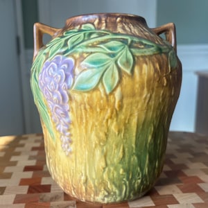 Op de afbeelding: Een keramische vaas met een getextureerde, schorsachtige buitenkant in tinten bruin, geel en groen. De vaas heeft een blauweregenontwerp met paarse bloemen en groene bladeren. Hij heeft twee kleine handvatten aan weerszijden.