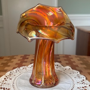 May include: An iridescent orange glass vase with a flared top and a twisted stem. The vase has a ruffled edge and a glossy finish, reflecting light with a rainbow sheen. The vase is a decorative item.