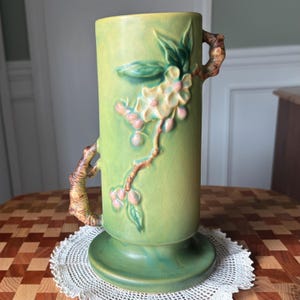 May include: A tall, green ceramic vase with a cylindrical shape and a wide base. The vase features a raised floral design with white flowers and pink berries. Two branch-like handles extend from the sides. The vase is approximately 10 inches tall.