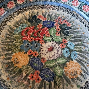 May include: A ceramic plate with a floral design. The plate features a central bouquet of flowers in shades of red, blue, orange, and white, surrounded by green leaves. The rim has a blue border with floral and scroll patterns.