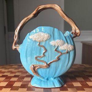 May include: A ceramic vase with a light blue body and a brown handle. The vase features a design of white clouds and brown tree branches. The handle is curved and resembles a tree branch. The vase is on a pedestal base.