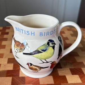 May include: A cream-colored ceramic pitcher featuring a series of colorful illustrations of various birds and the text "BRITISH BIRDS" printed in light blue letters near the top rim. The pitcher has a curved handle and a wide pouring spout.