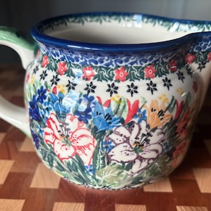 May include: A ceramic pitcher with a floral design. The pitcher features a white base with a colorful pattern of flowers in red, blue, yellow, and white. It has a blue rim and a green handle.