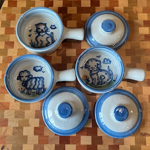 May include: Three ceramic bowls with handles and matching lids, featuring hand-painted blue designs of a cat, a sheep, and a dog on the interior base. The bowls have a white glaze with blue rim accents and a smooth, rounded shape for serving.