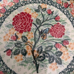 May include: A ceramic plate with a floral design. The plate features a central motif of red roses and a butterfly, surrounded by smaller orange flowers and green leaves. The rim has a blue and white pattern.