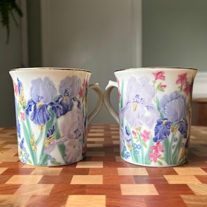 May include: Two white ceramic mugs featuring a floral pattern of purple and blue iris flowers with green stems and small pink blossoms. Each mug has a gold-trimmed rim and a curved handle, displaying a detailed botanical design on the exterior.
