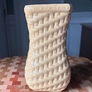 May include: A cream-colored ceramic vase with a woven basket design. The vase has a slightly flared shape and a textured surface, giving it a rustic appearance. The vase is sitting on a wooden surface.