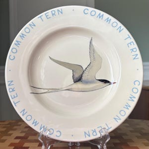 May include: A round ceramic plate with a cream-colored surface featuring a central illustration of a common tern bird in flight. The rim of the plate is decorated with the repeating light blue text "COMMON TERN" separated by small dots.