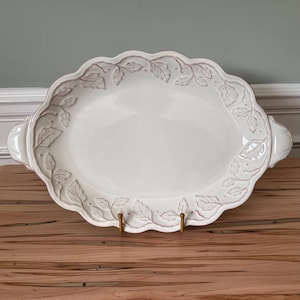 May include: A white oval serving platter with a scalloped edge and embossed leaf design. The platter has two handles and is displayed on a wooden surface. The leaf design is accented with a light brown color.
