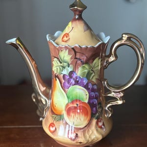 May include: A decorative ceramic teapot with a gold handle, spout, and lid finial. The teapot is painted with a colorful fruit design featuring grapes, apples, pears, and cherries. The background is a light brown color.