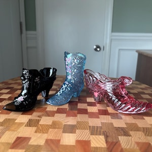 May include: Three decorative glass shoe figurines. One is black with floral accents, another is a light blue boot with butterfly details, and the third is a pink high-heeled shoe. All have intricate designs.