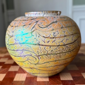 May include: A spherical glass vase featuring a swirling pattern of yellow, orange, and brown hues with an iridescent finish. The surface displays a complex web of fine lines and organic curves that wrap around the rounded body of the vessel.