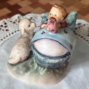 May include: A ceramic figurine depicting a child holding a pink object, seated in a blue and green cradle, with a lamb beside them. The cradle has red floral accents. The figurine is on a green base.