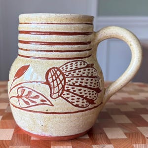 May include: A ceramic mug with a speckled cream glaze featuring hand-painted reddish-brown horizontal stripes around the upper rim and a large, detailed floral motif on the body, complete with a curved handle and a solid reddish-brown base.