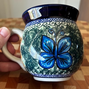 May include: A ceramic mug with a blue butterfly design. The mug features a dark blue rim and handle, with a white and green floral pattern. The butterfly is a vibrant blue with black outlines. The mug is likely hand-painted.
