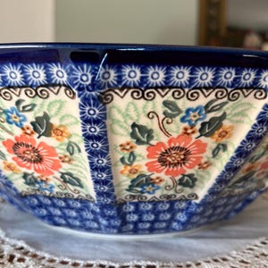 May include: A ceramic bowl with a vibrant floral pattern. The bowl features a deep blue rim and base, with panels of white and light green adorned with orange, blue, and yellow flowers. The design includes intricate scrollwork and star-like accents.