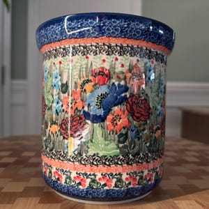 May include: A ceramic utensil holder with a vibrant floral design. The cylindrical holder features a blue rim and base, with a band of orange and black detailing. The body is covered in colorful flowers, including red, blue, and orange blooms.
