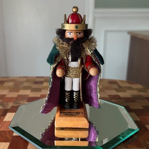 May include: A wooden nutcracker in the shape of a king, wearing a red and gold crown, green cape, and gold accents. The nutcracker is standing on a mirrored surface, with the text "Tchaikovsky's 'Prince'" on the base.