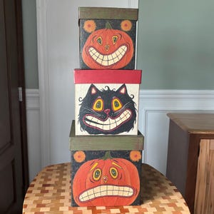 May include: A stack of three square boxes with lids. The top box features an orange pumpkin with a wide grin on a black background. The middle box shows a black cat with yellow eyes on a cream background. The bottom box displays an orange pumpkin.