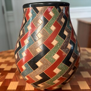 May include: A bulbous ceramic vase featuring a complex geometric pattern of interlocking lines. The surface is decorated with a repeating motif in shades of red, green, black, and speckled tan, finished with a solid black rim at the top opening.