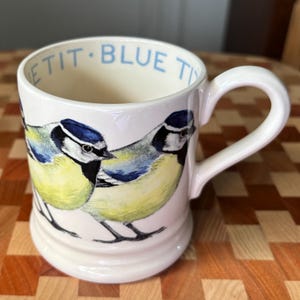 May include: A cream-colored ceramic mug featuring a detailed illustration of two blue tits with yellow chests, blue caps, and black eye stripes. The interior rim of the mug displays the text BLUE TIT in light blue lettering on a cream background.
