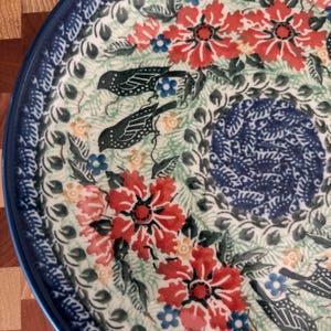 May include: Round ceramic plate with a blue rim and a floral design. The plate features red flowers, blue accents, and green leaves. Two black bird illustrations are also included in the design. The plate has a white background.