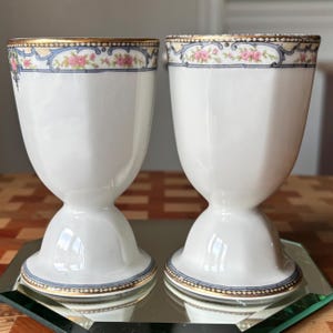 May include: Two white porcelain egg cups with a gold rim and a decorative floral and blue border. The egg cups have a pedestal base and a flared top. The floral design includes pink roses.