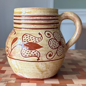 May include: A ceramic mug with a rounded body and a single handle, featuring a speckled yellow and tan glaze. The exterior is decorated with hand-painted brown floral and leaf motifs, with horizontal bands encircling the upper rim area.