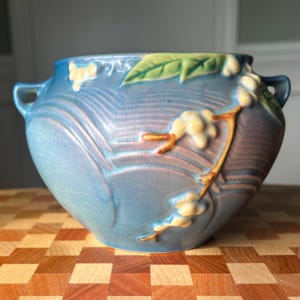 May include: A blue ceramic bowl with two handles and a floral design. The bowl features a raised pattern and is decorated with a branch of white flowers and green leaves. The bowl is sitting on a wooden surface.