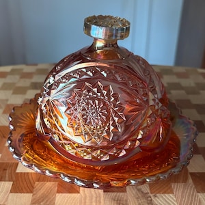 May include: An amber-colored iridescent glass butter dish with a domed lid and a matching base. The glass features a pressed geometric starburst pattern with diamond-shaped facets and a scalloped edge on the base, topped with a round knob.